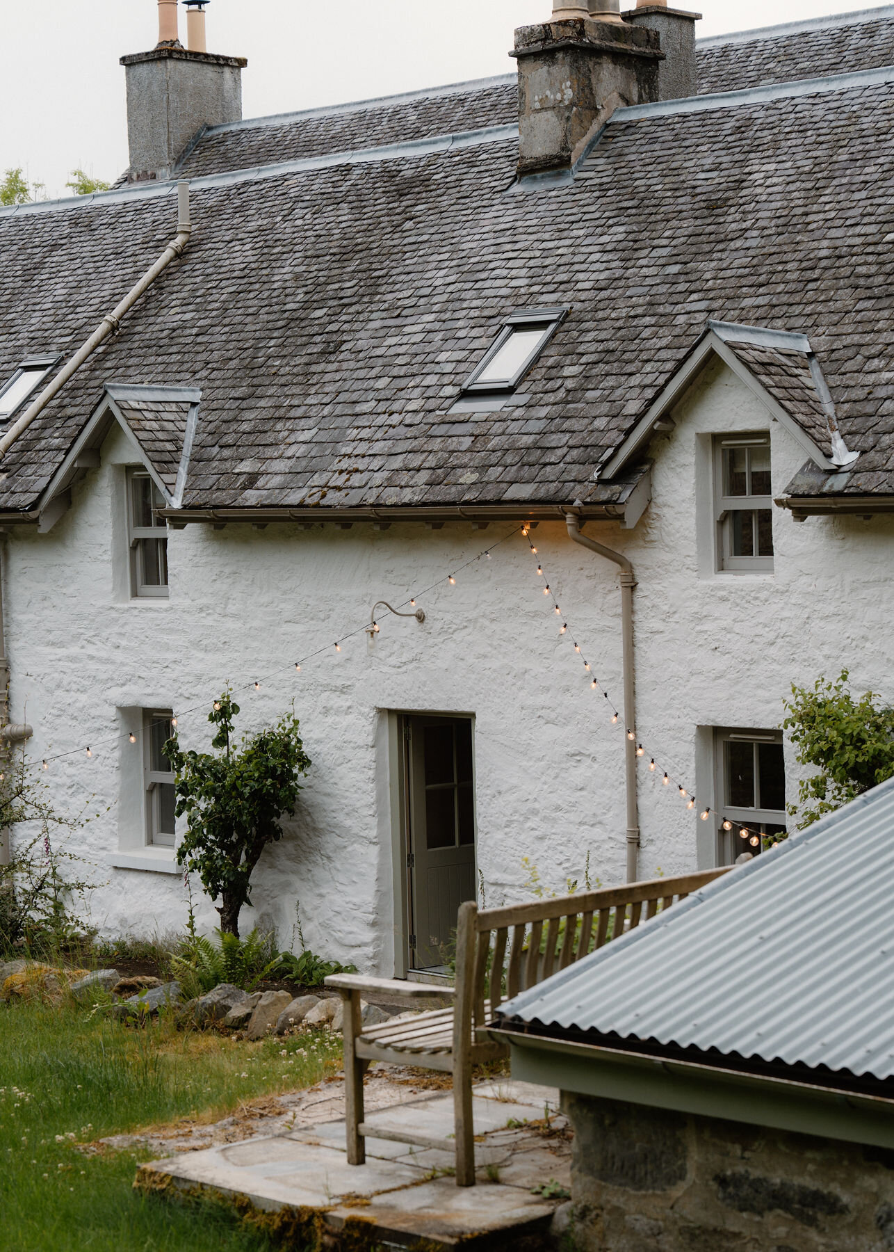 The back of a white harled cottage with slate roof and dormer windows, festoon lights across the wall and a teak bench in front.