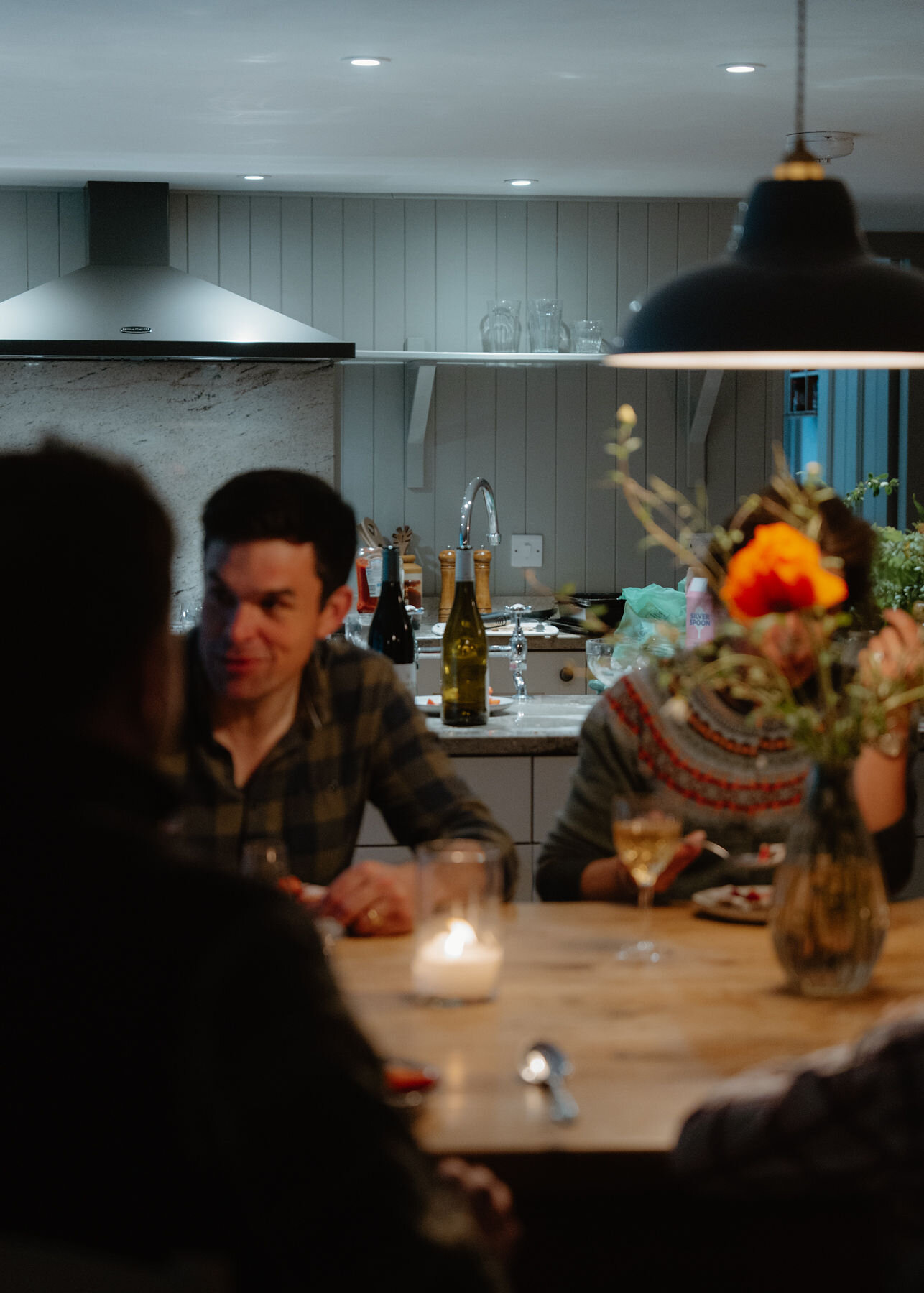 A man in a checked shirt talks across the candlelit table, beside an orange poppy and a woman in a Fair Isle cardigan with a glass of wine.