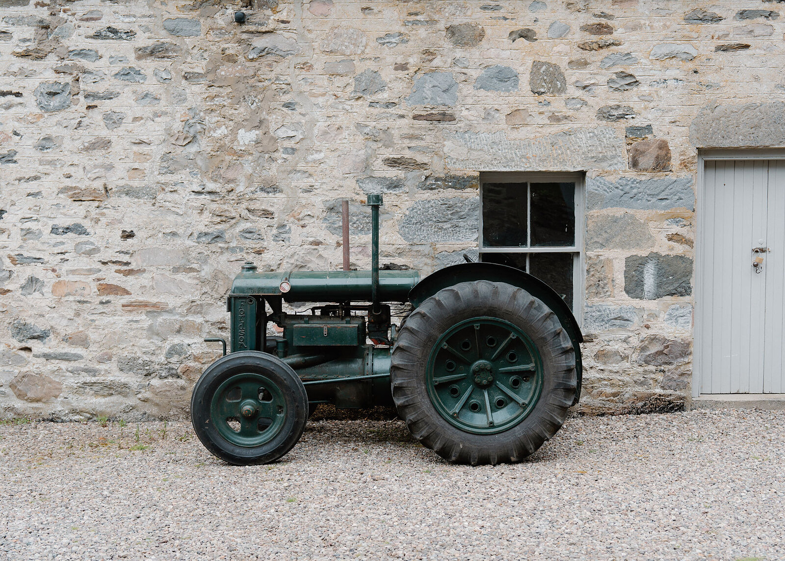 The vintage green tractor in a wider landscape frame on gravel against the stone steading wall, a sash window and pale grey door beside it.