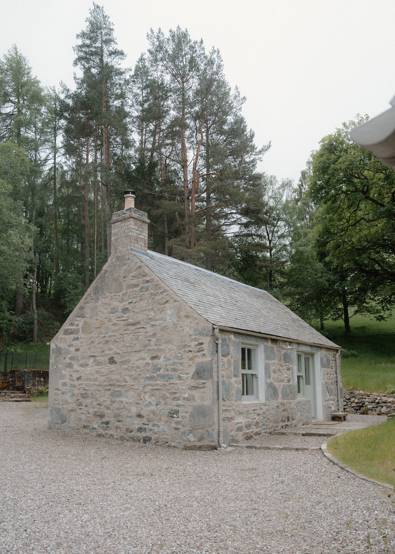 A small stone bothy with a slate roof and a single chimney stands on gravel beneath tall Scots pines, seen from the gable end.