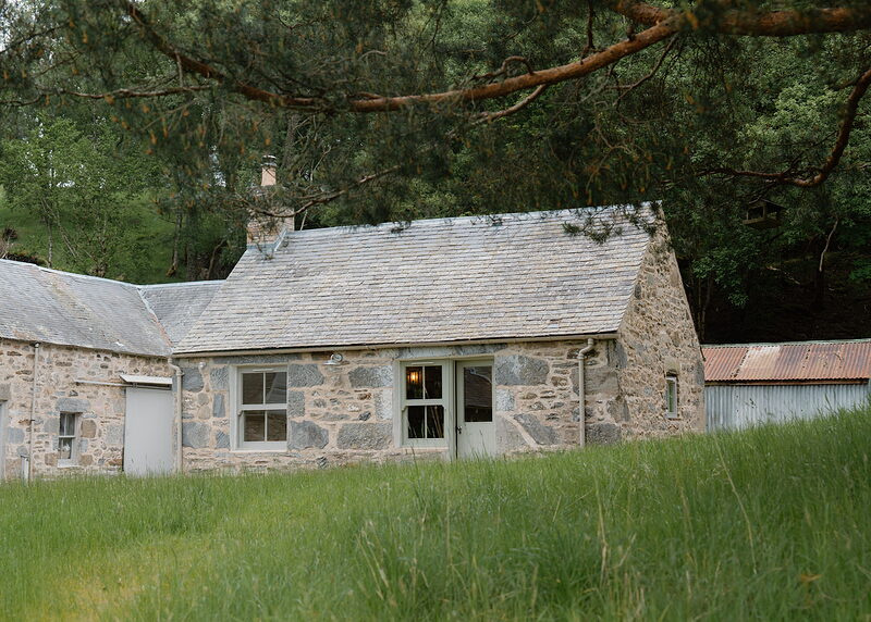 A small stone bothy joined to a stone steading, seen across long grass under a Scots pine bough, a corrugated shed to the right.