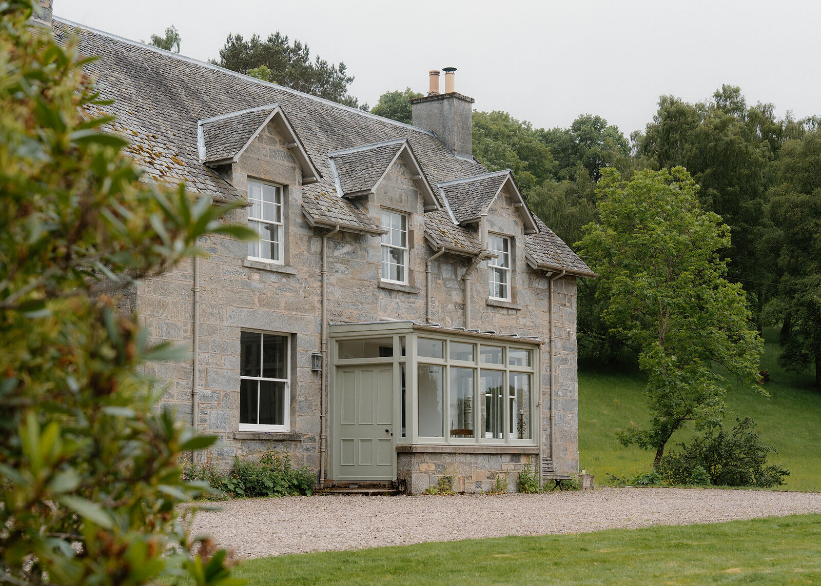 A landscape view of the grey stone house with its dormers and glazed porch, a birch tree and wooded hillside to the right.
