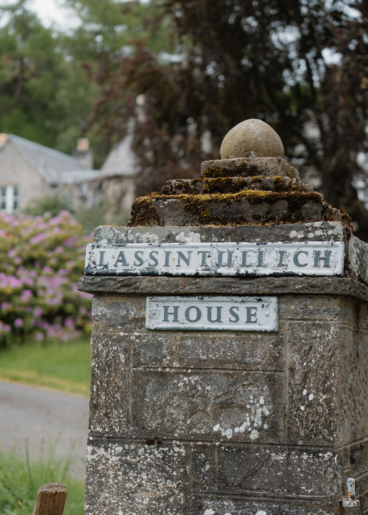 Close on the mossy stone gate pier and its chipped plaques reading LASSINTULLICH HOUSE, rhododendrons and the house blurred behind.