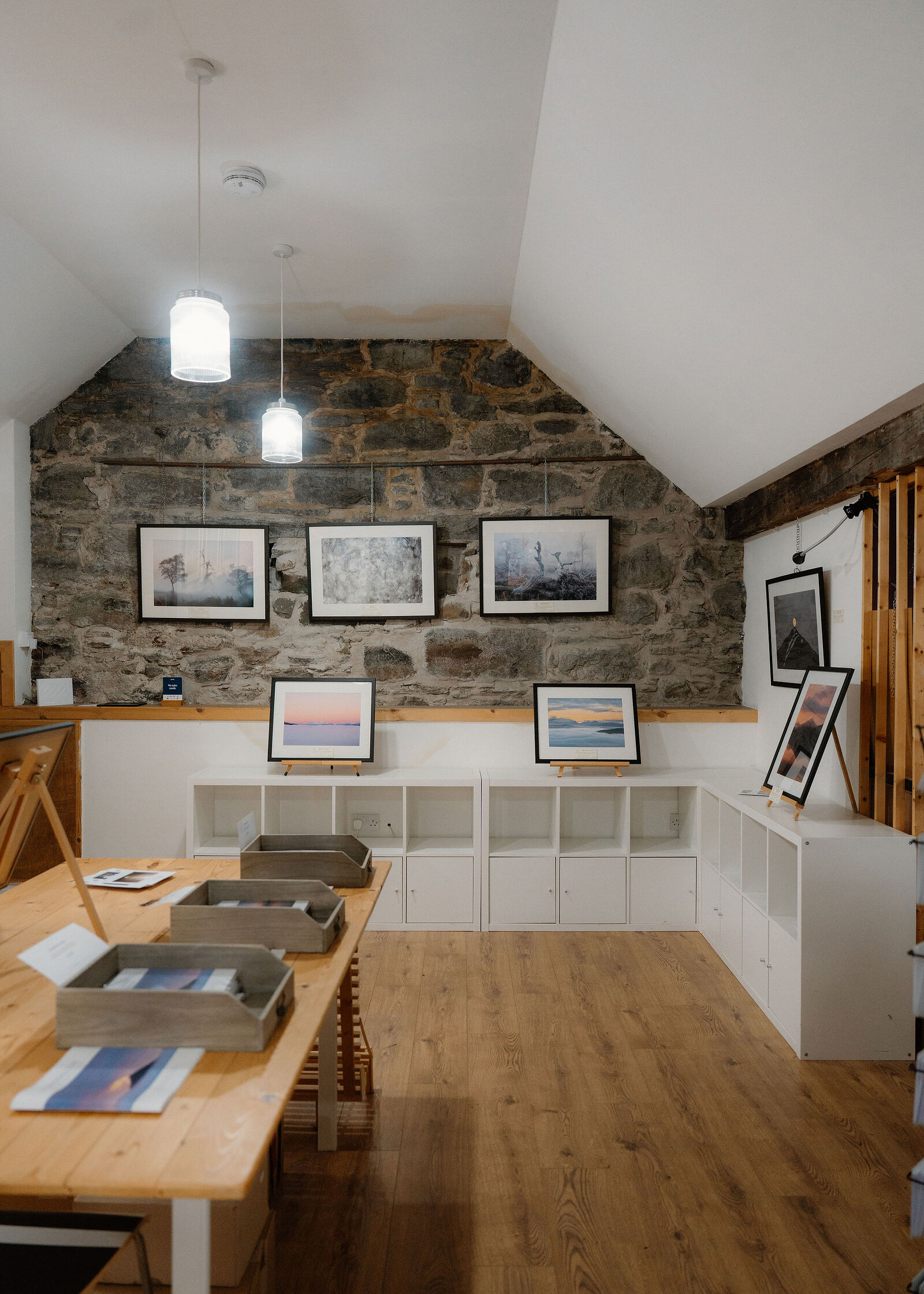 Inside a small gallery, framed landscape photographs hang on a stone gable wall under pendant lamps above white shelving and a pine table.