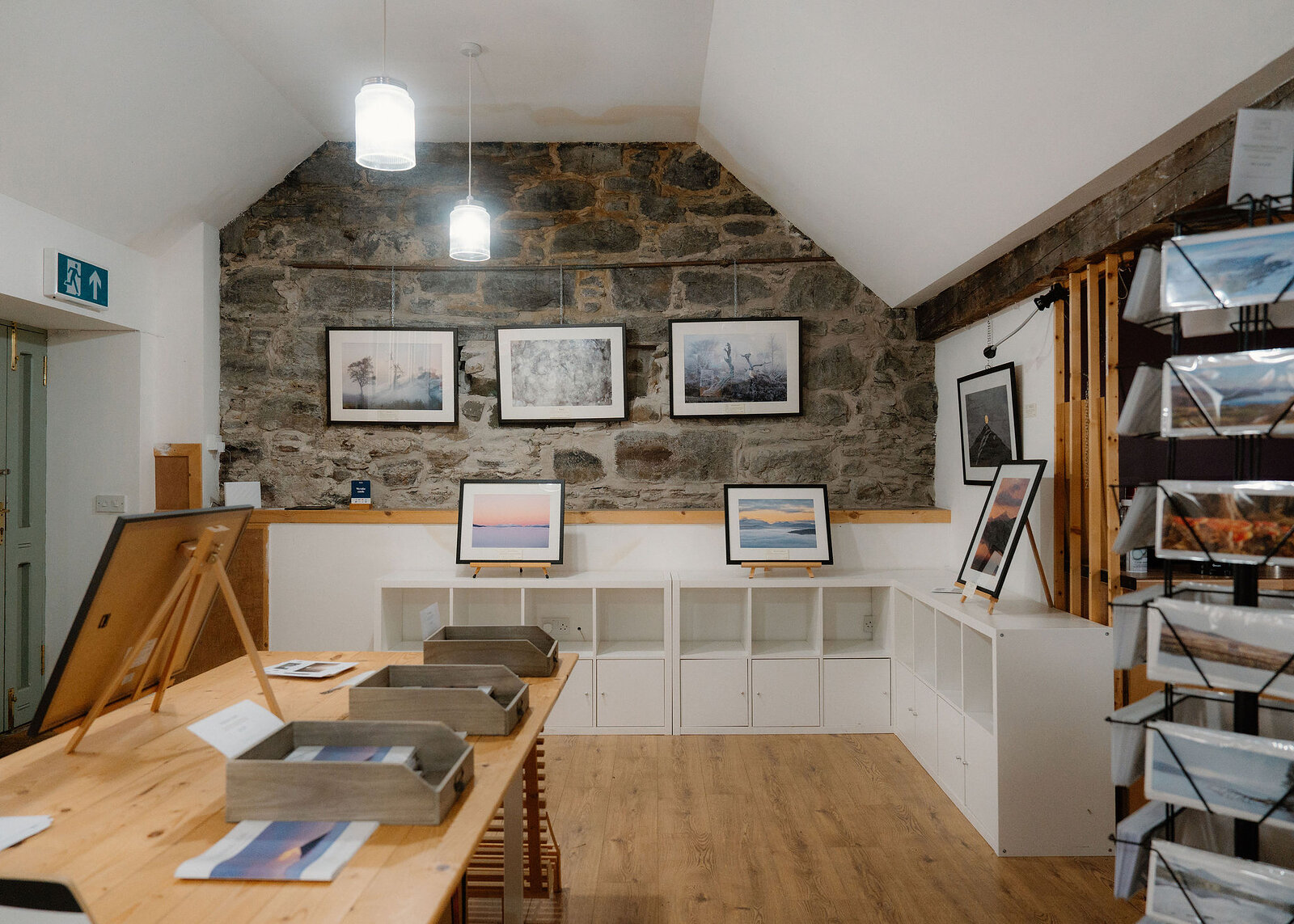 A wide view of the gallery room, with the green door and exit sign on the left and a postcard rack on the right.