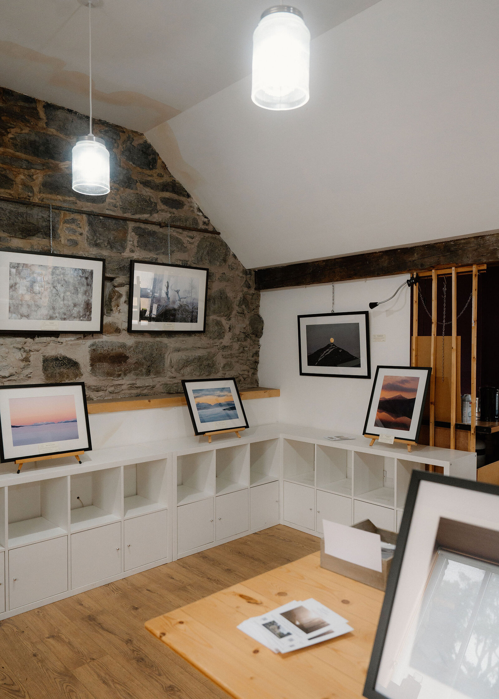 A gallery corner with a pendant lamp close by and framed prints on easels, including a full moon above a pointed peak.
