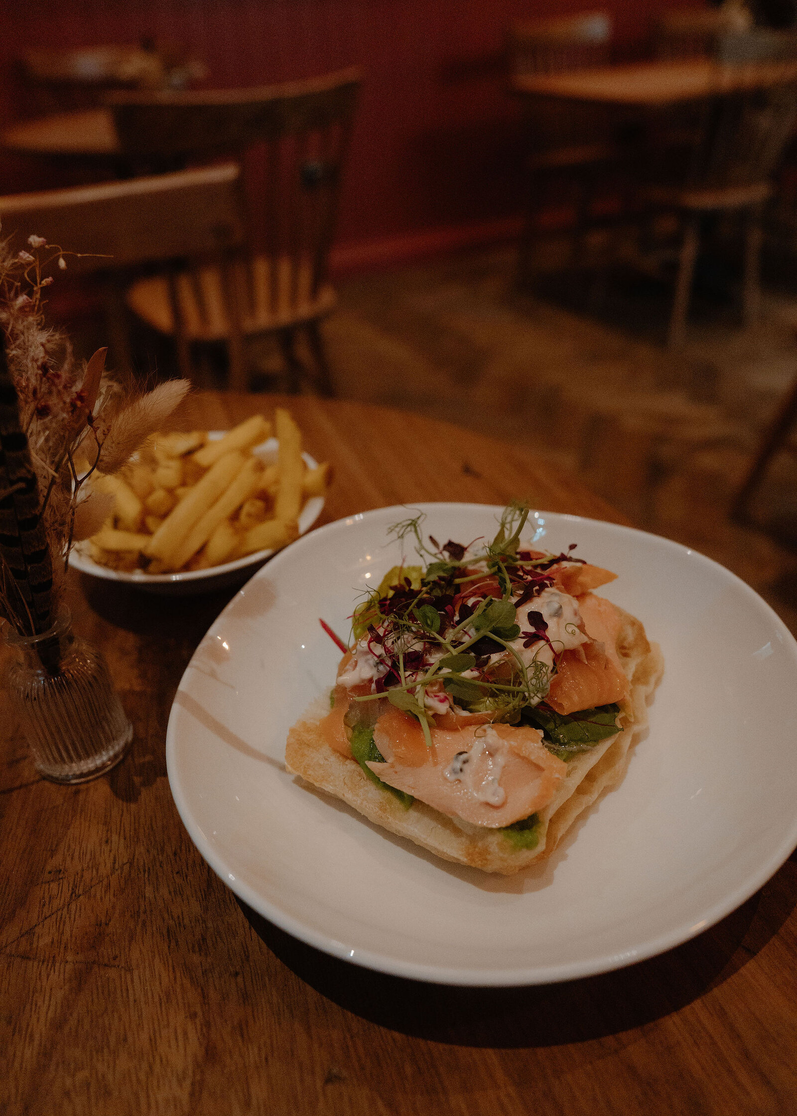 Smoked salmon on toasted bread with microgreens and dressing sits on a white plate beside a bowl of chips and dried flowers.