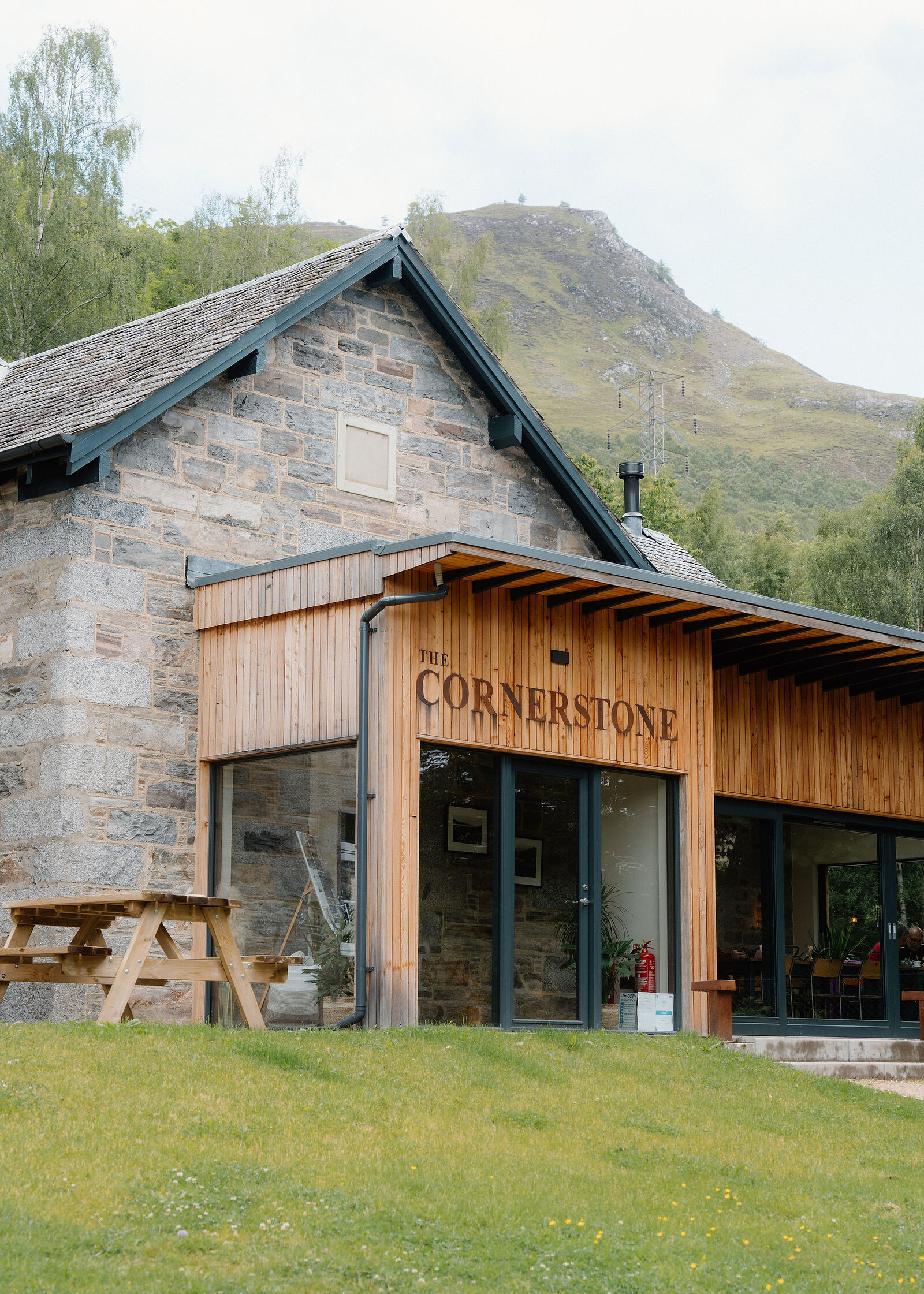The Cornerstone lettering on larch cladding beside a stone gable, a picnic bench on the lawn and the craggy hill behind.