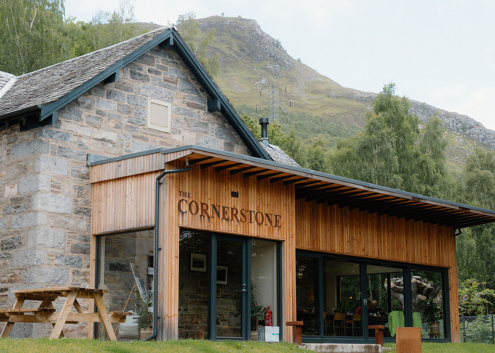 A wider view of The Cornerstone's stone and larch building, diners visible through its glass, with the hill and pylon behind.