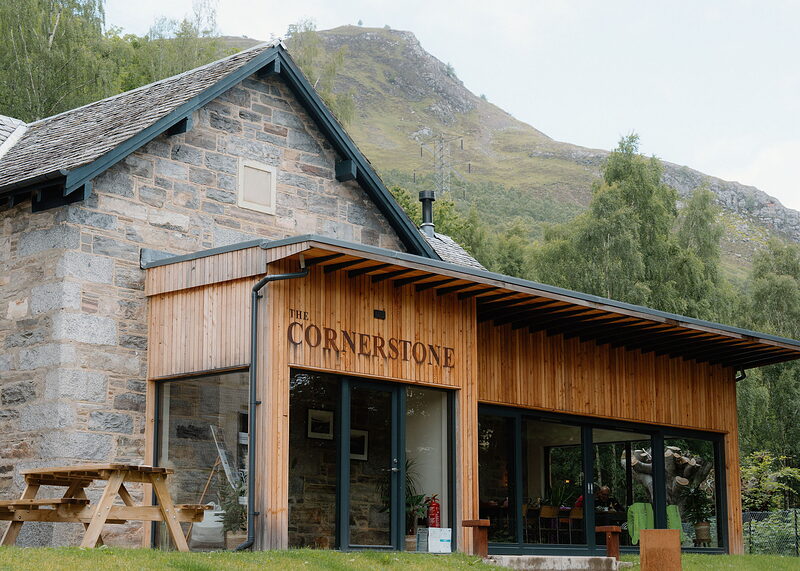 A wider view of The Cornerstone's stone and larch building, diners visible through its glass, with the hill and pylon behind.