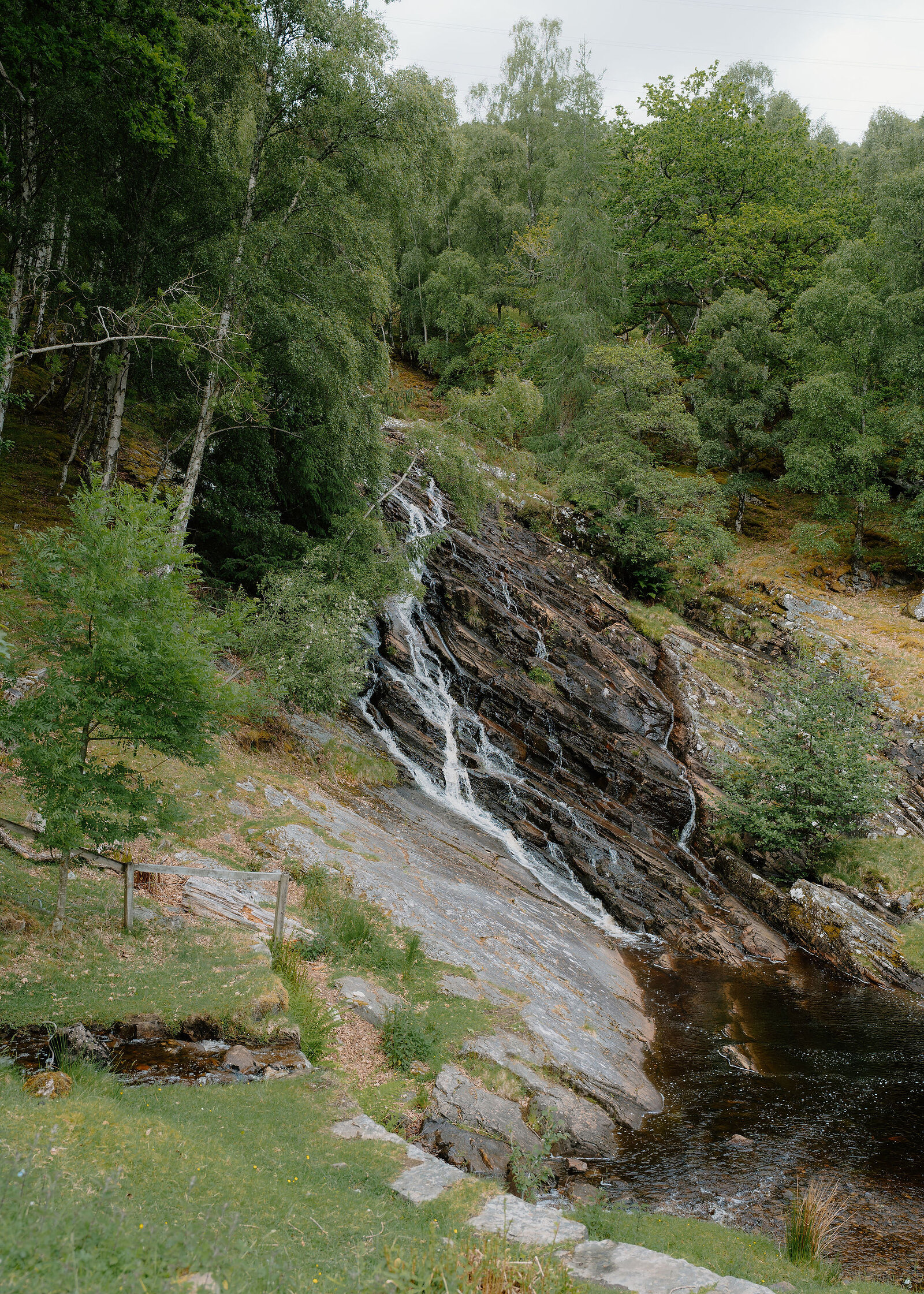 A small waterfall pours over tilted dark rock slabs into a brown pool, with birch woodland and a low wooden fence around.