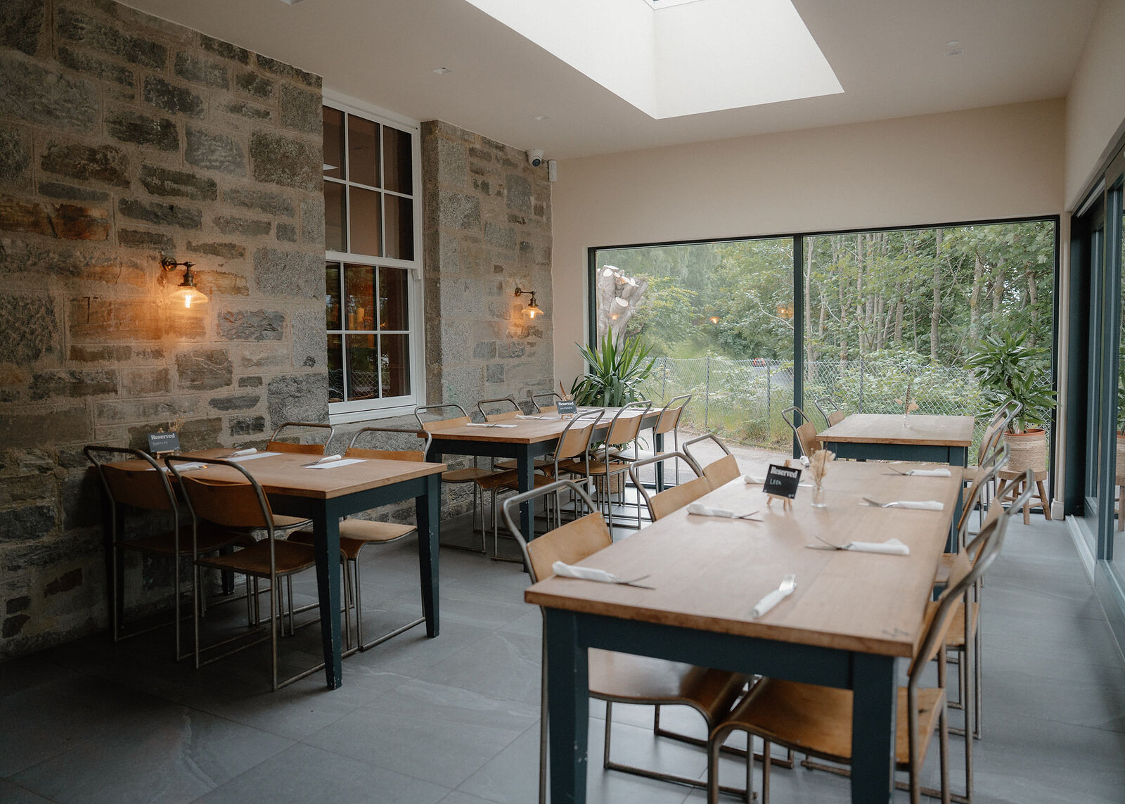 A wider landscape view of the rooflit dining room, more tables and chairs, a big window onto trees and potted plants.