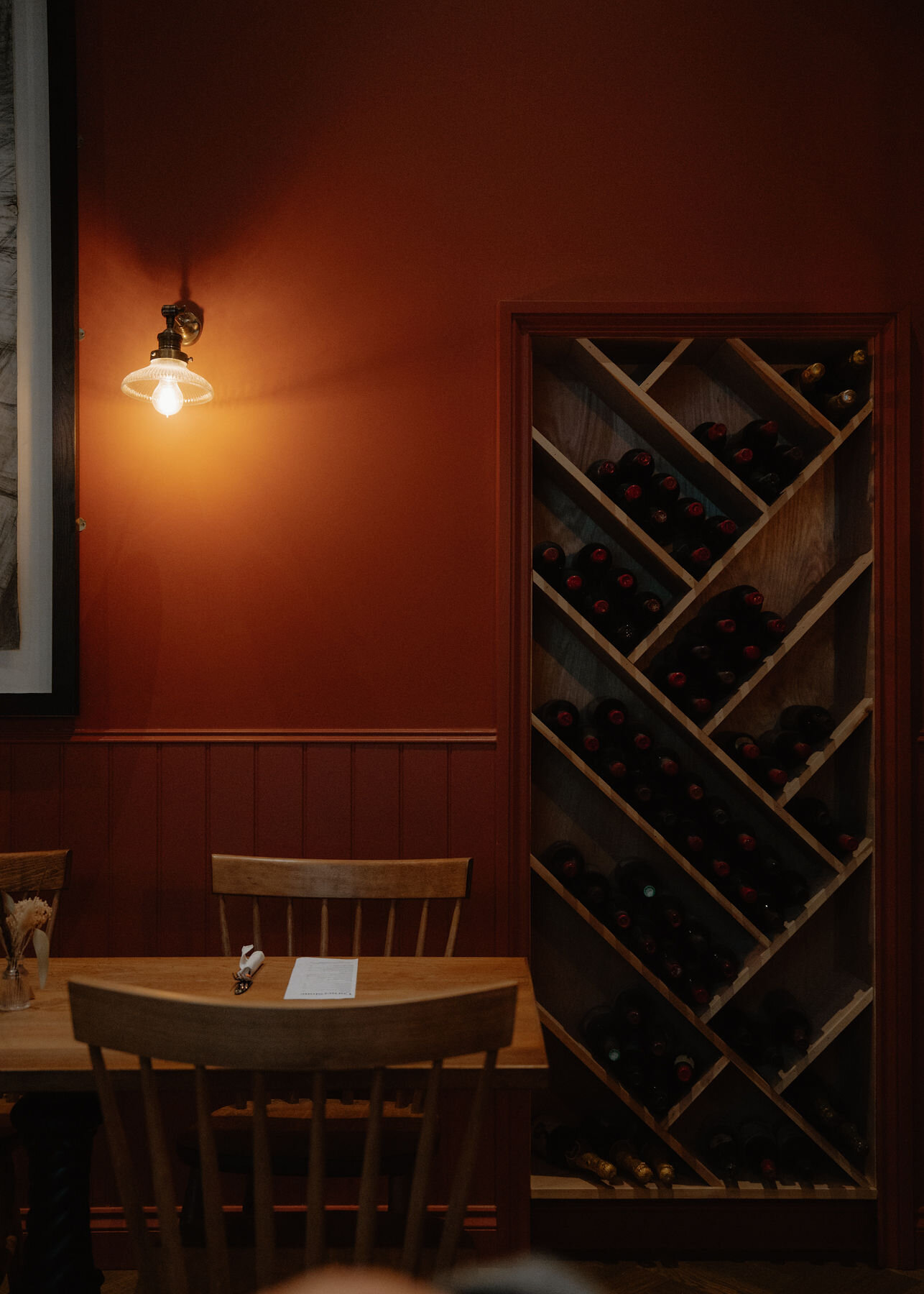 A diagonal wine rack in a terracotta wall beside a lit brass lamp, a laid table in front and a blurred head below.