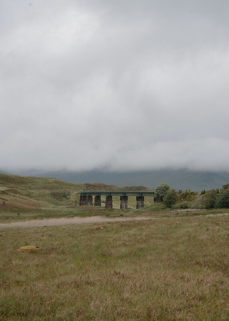 A green lattice railway viaduct on dark stone piers crosses a moorland hollow, with low cloud hiding the hills and rough grass in front.