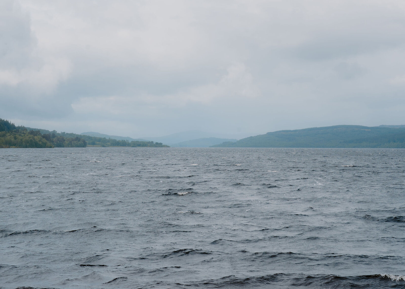 Choppy dark water fills most of the frame, the loch running away between forested slopes to distant hills in haze.