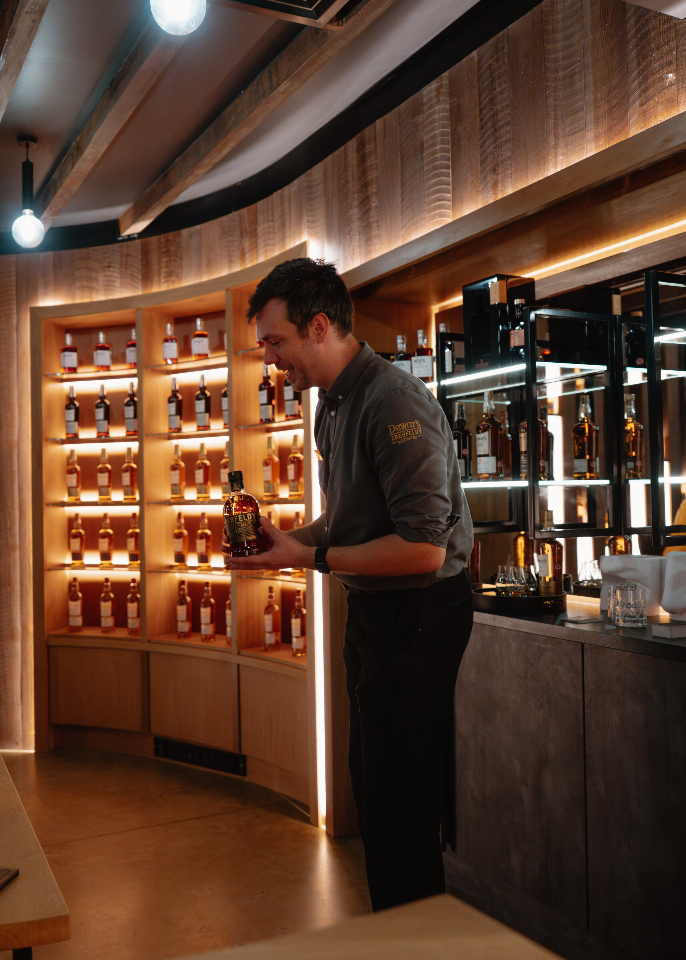 A host in a Dewar's shirt takes a bottle from the backlit shelves of the tasting room.