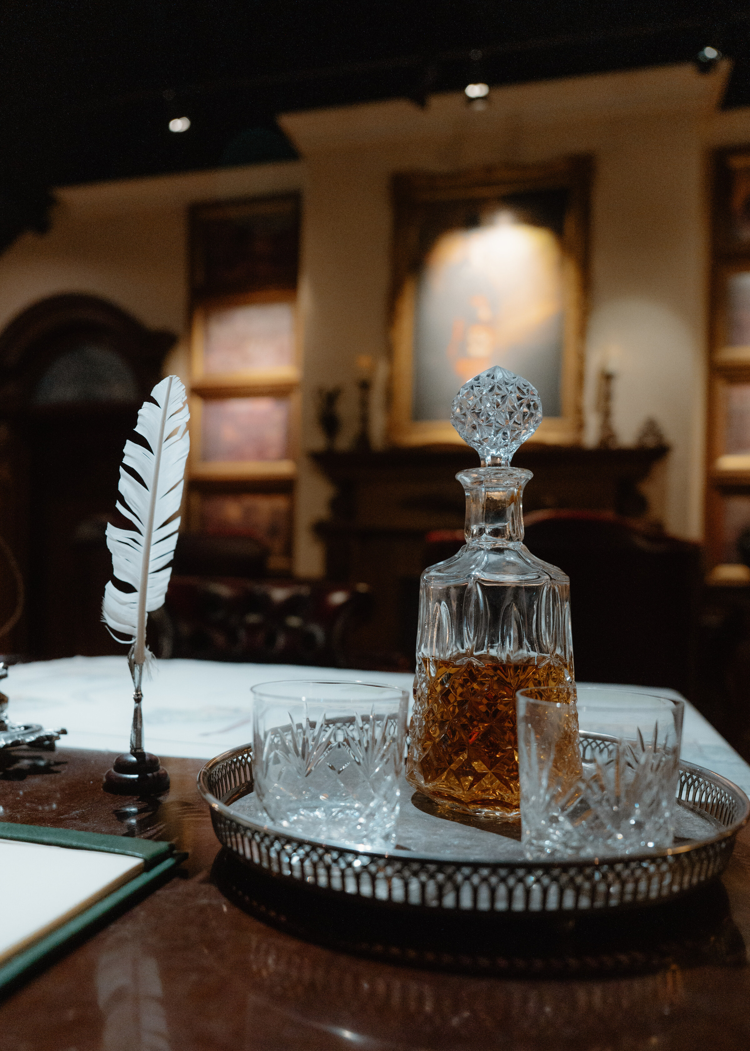 A cut-glass decanter of whisky and two tumblers on a silver tray beside a white quill, on a polished desk in the museum.
