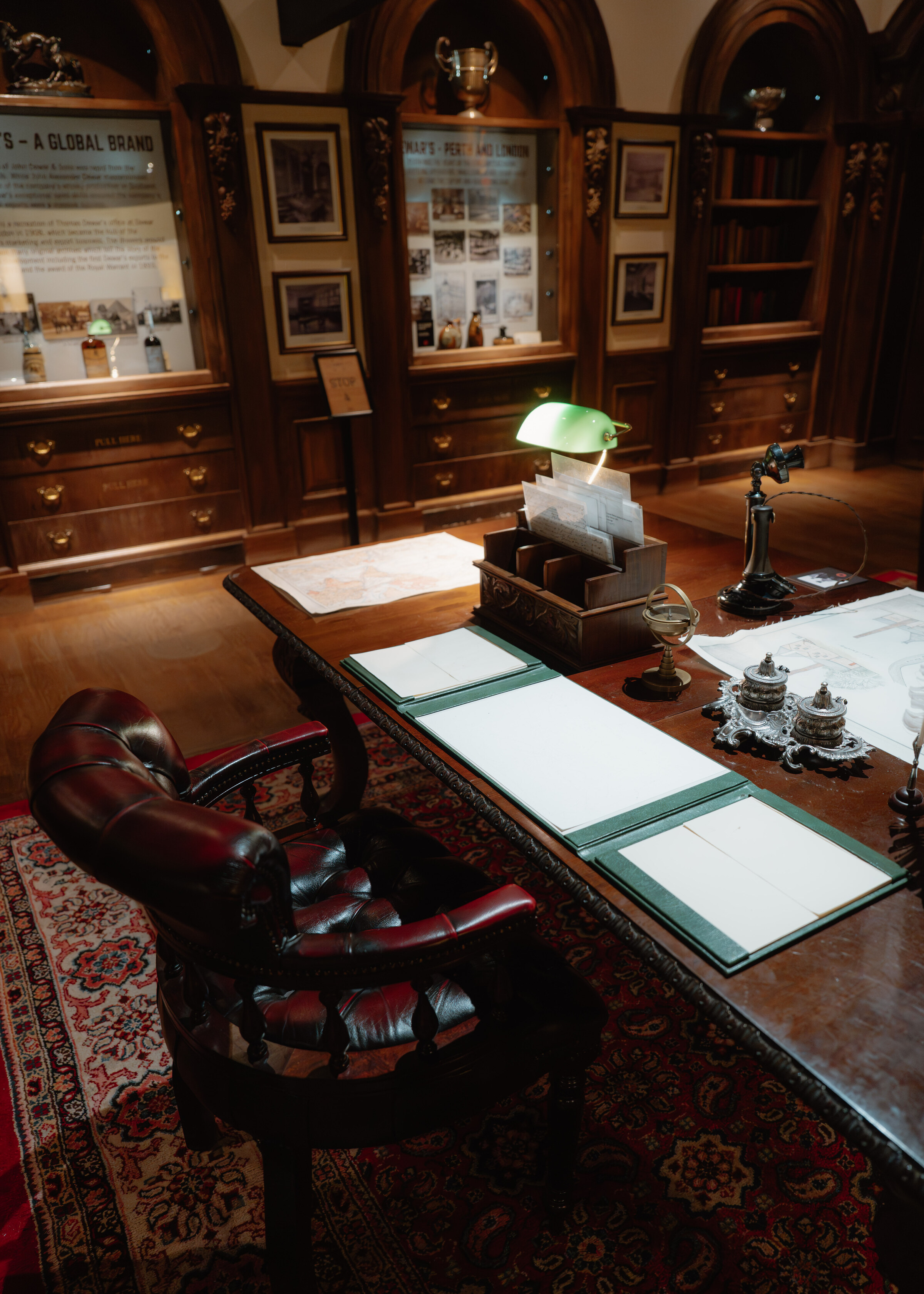 A Victorian desk with a green-shaded lamp, ledgers and a leather chair in the museum, glass cases and framed papers behind.
