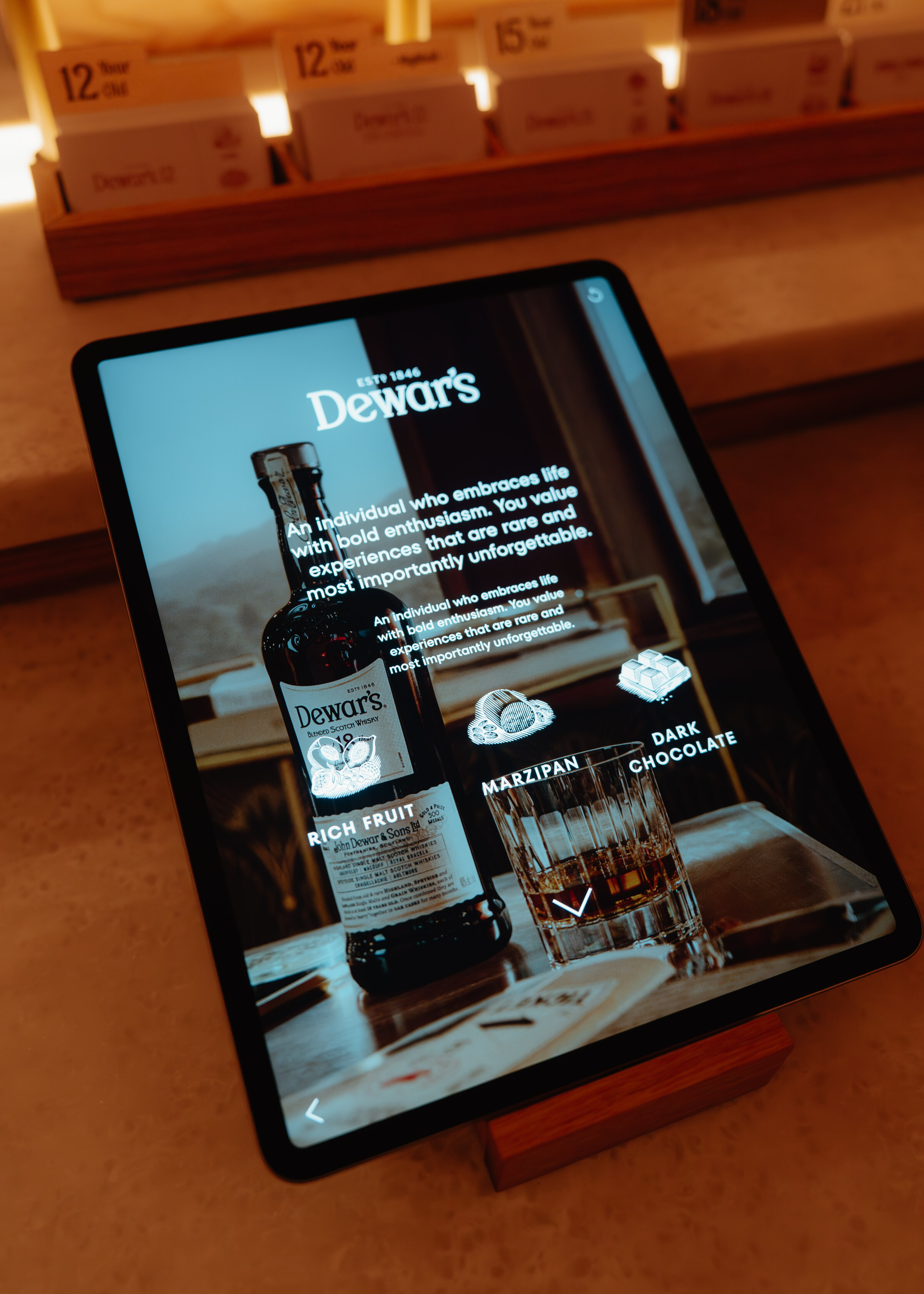 A tablet on a wooden stand showing a Dewar's bottle and its tasting notes, rich fruit, marzipan and dark chocolate.