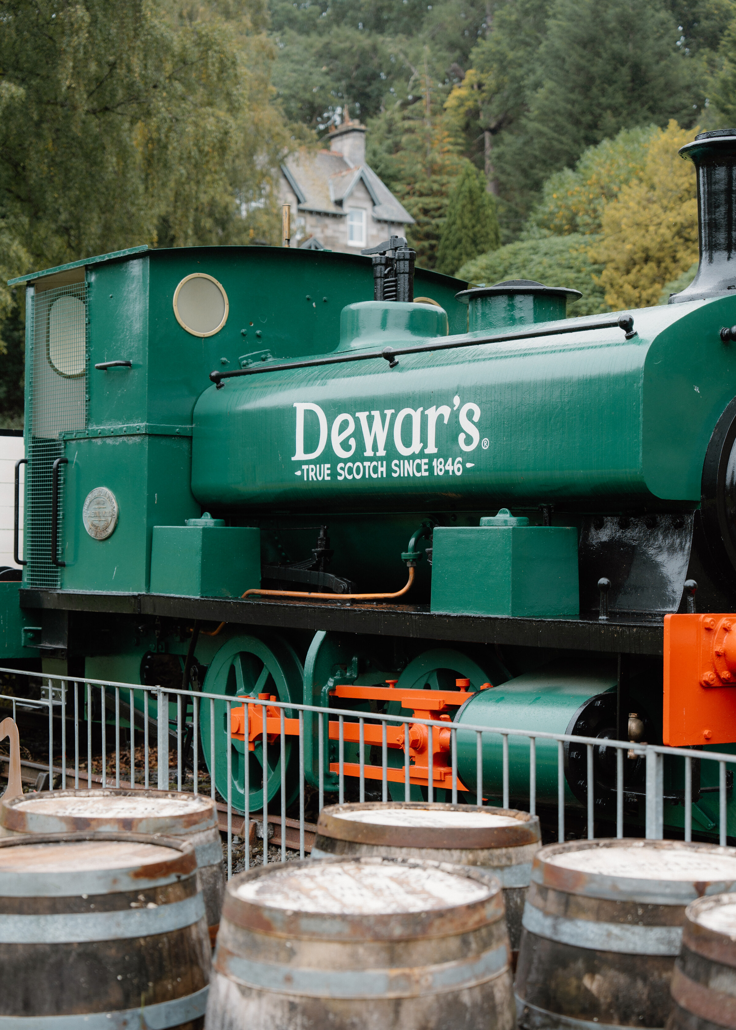 A green tank engine painted Dewar's, True Scotch since 1846, behind a rail with casks in front of it and woodland beyond.