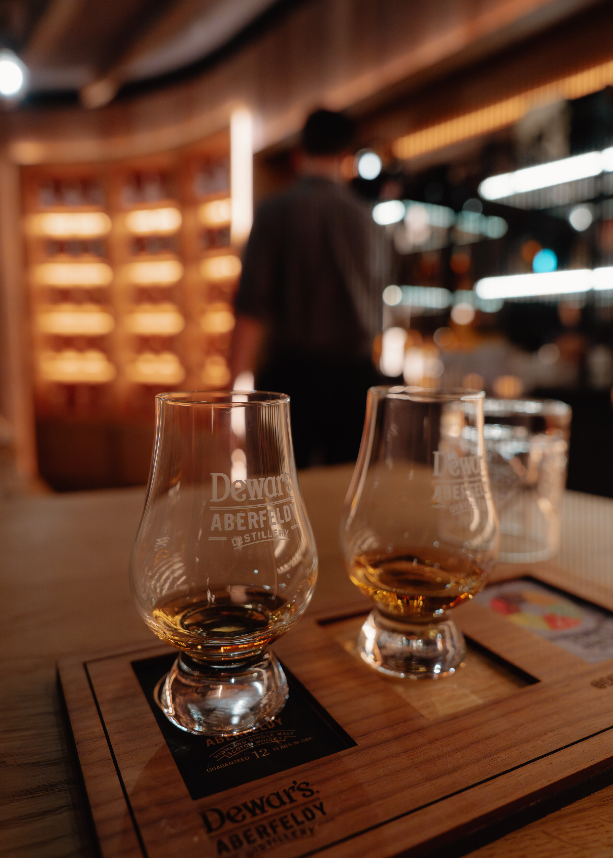 Two Glencairn glasses of whisky on a tasting board marked Dewar's Aberfeldy, the lit shelves of the tasting room behind.