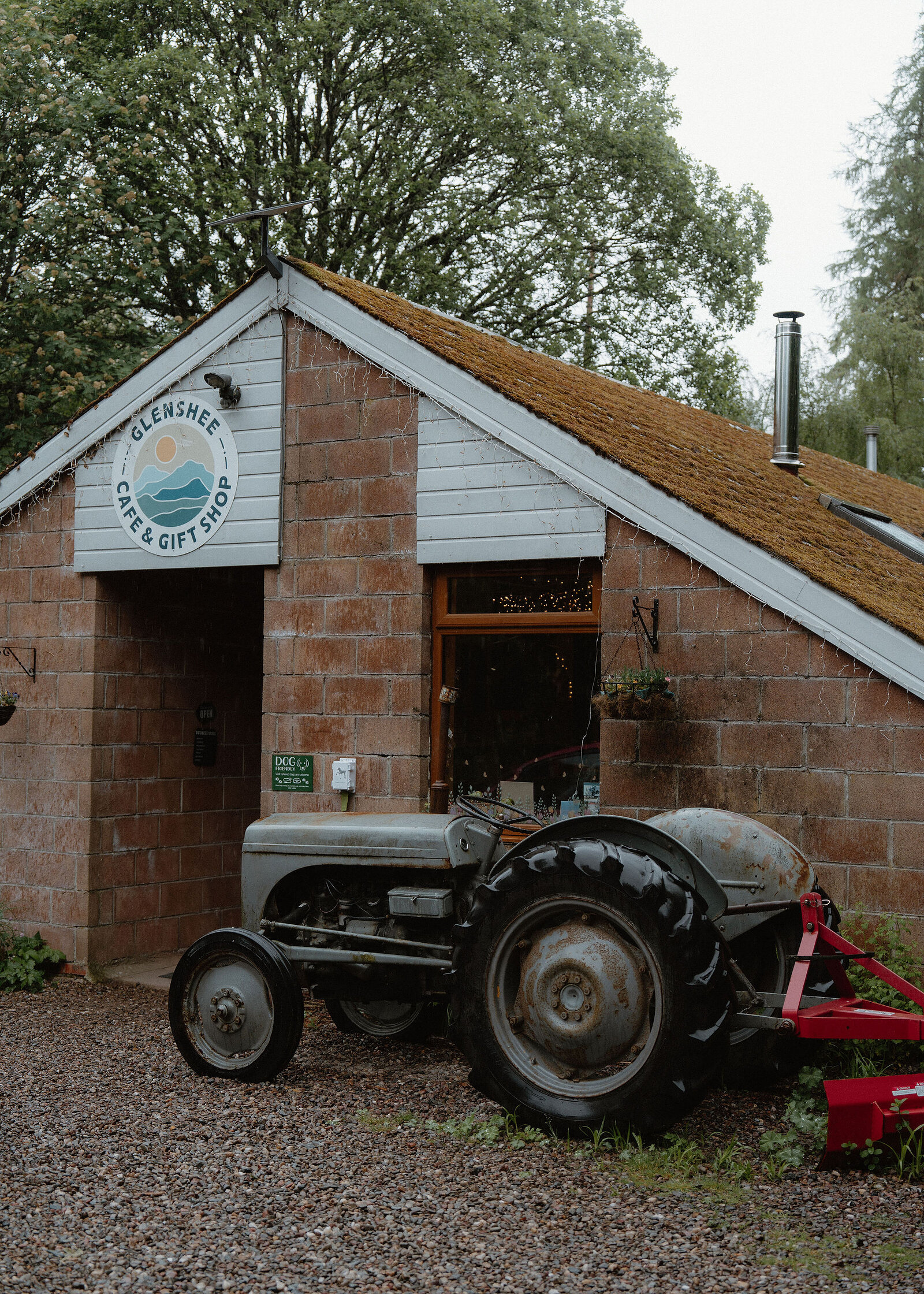A grey vintage tractor with a red implement stands on the gravel outside the brick Glenshee Cafe and Gift Shop, its round sign on the gable.