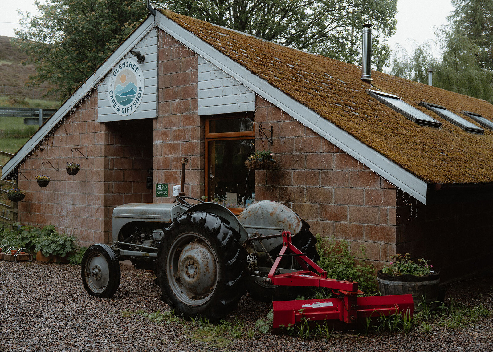The Glenshee Cafe and Gift Shop, a brick building with a mossy roof and roof lights, a grey tractor parked outside and the hill behind.