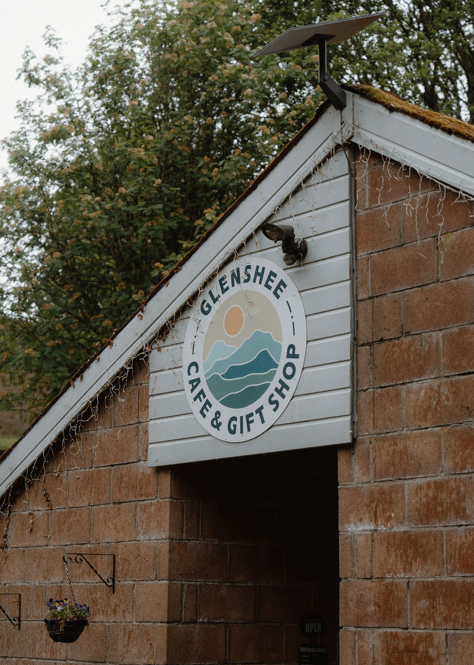 The round Glenshee Cafe and Gift Shop sign, hills and a sun, on the white-boarded gable above the door.
