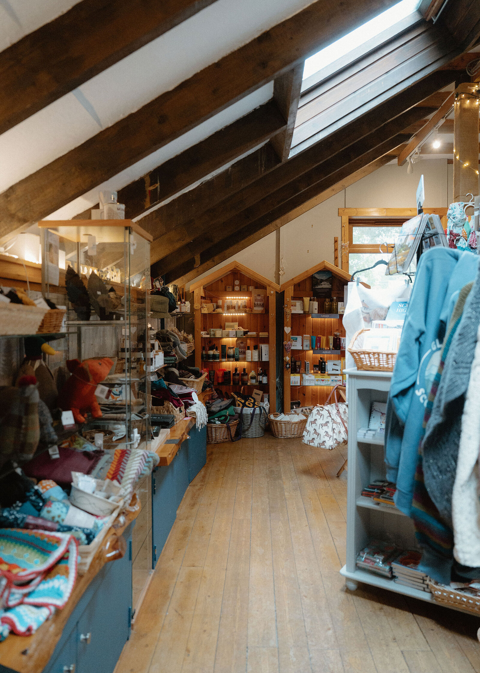 The gift shop under exposed beams and a roof light, shelves of knitwear, crafts and gifts running down a wooden floor.