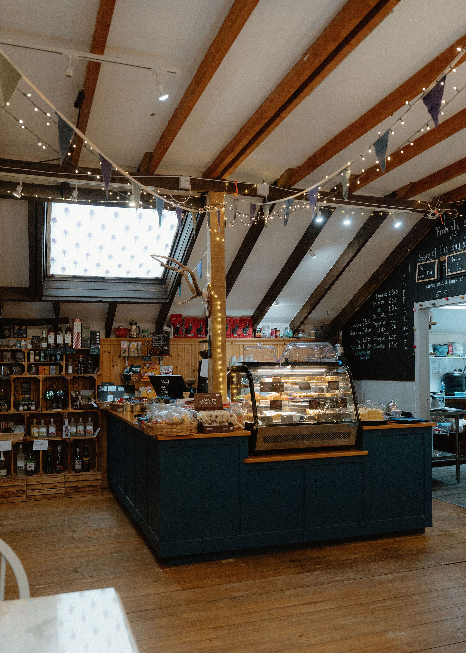 Inside the Glenshee Cafe: a dark blue counter with a glass cake cabinet under timber beams, bunting and fairy lights, shelves of bottles behind.