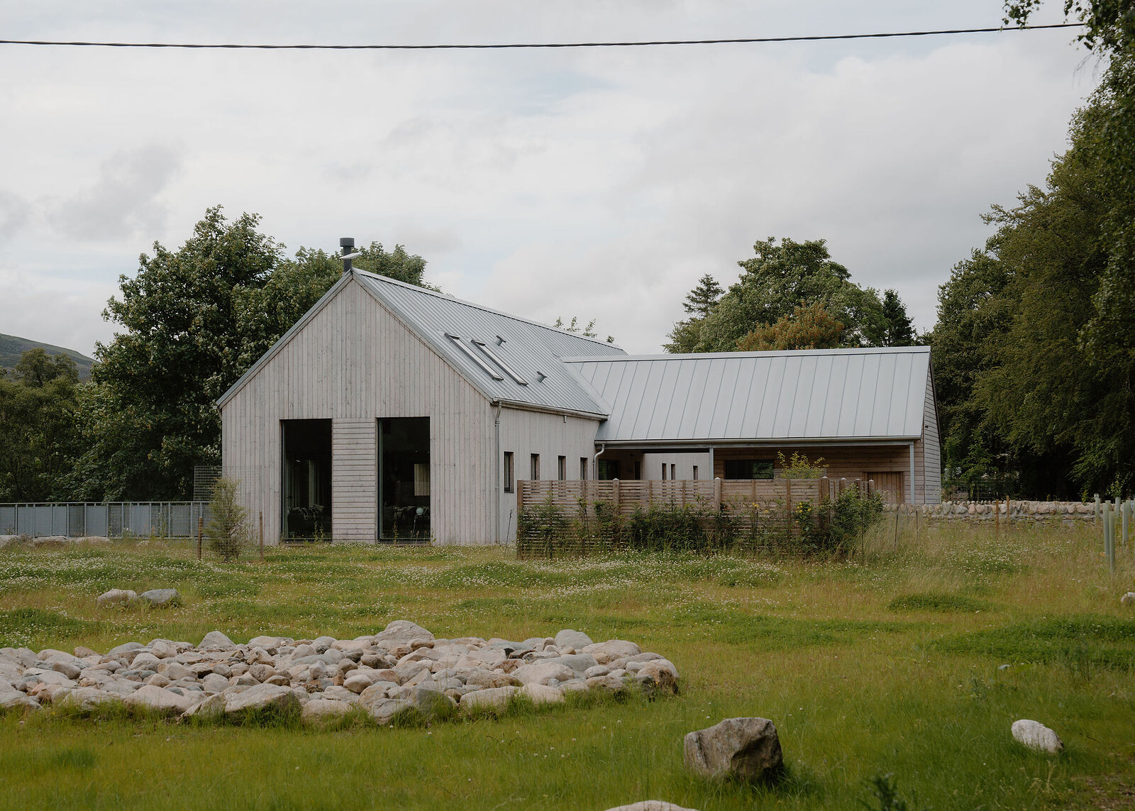 The whole of Larch House across its meadow: the larch-clad gable and long roof with roof lights, the lower wing beside it, a ring of river stones in the grass and oaks behind.