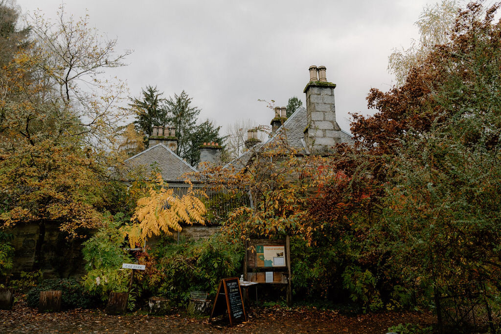 A grand stone mansion with clustered chimneys glimpsed through autumn foliage, an information board set beside the path