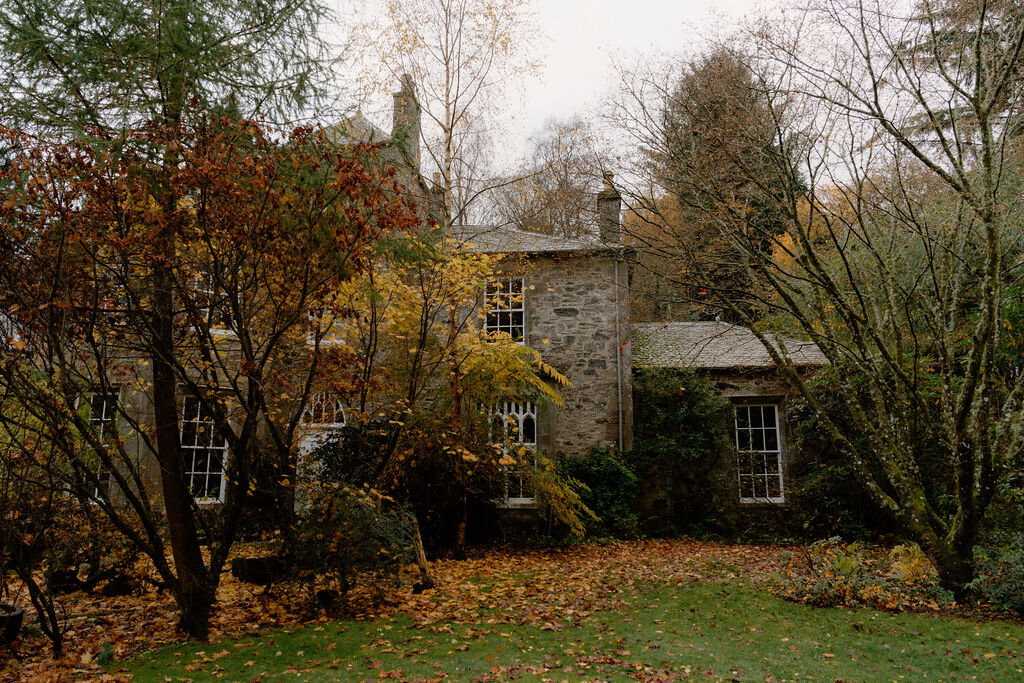 A stone mansion glimpsed through autumn trees, fallen leaves scattered across the lawn