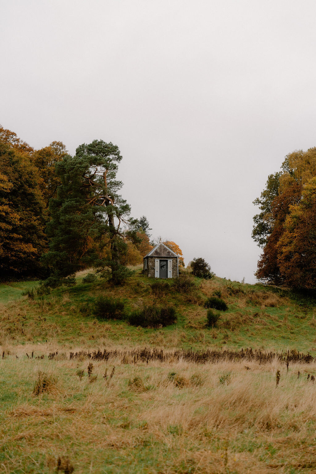 A stone folly with a pointed slate roof crowns a grassy knoll circled by autumn trees.