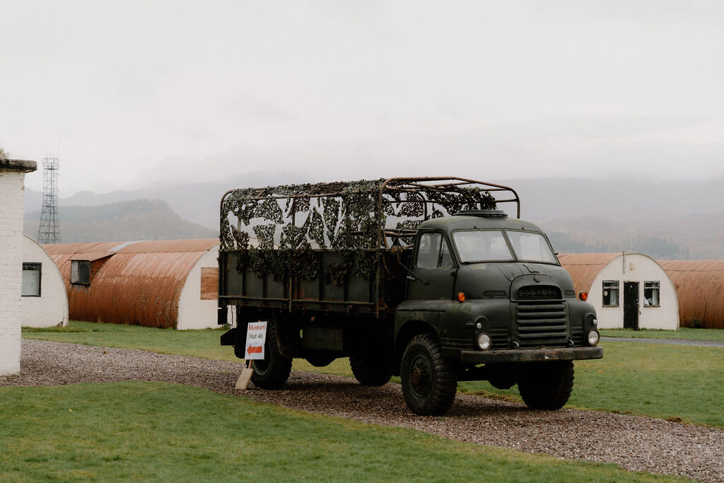 A vintage military truck stands parked outside a row of corrugated Nissen huts.