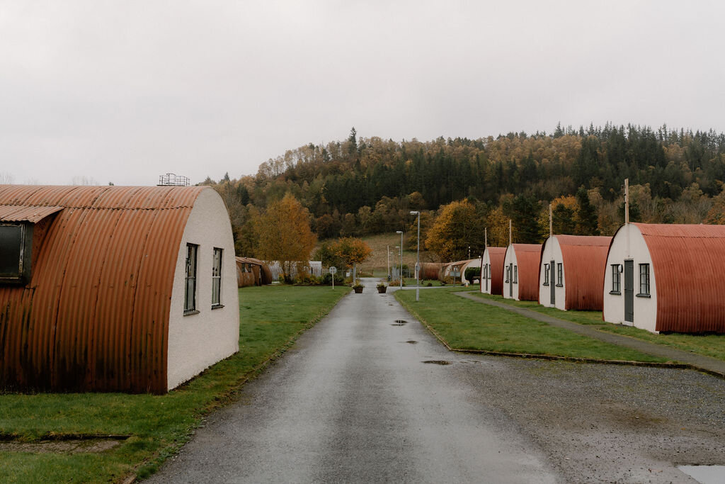 A paved road runs between rows of corrugated Nissen-hut chalets beneath autumn trees.