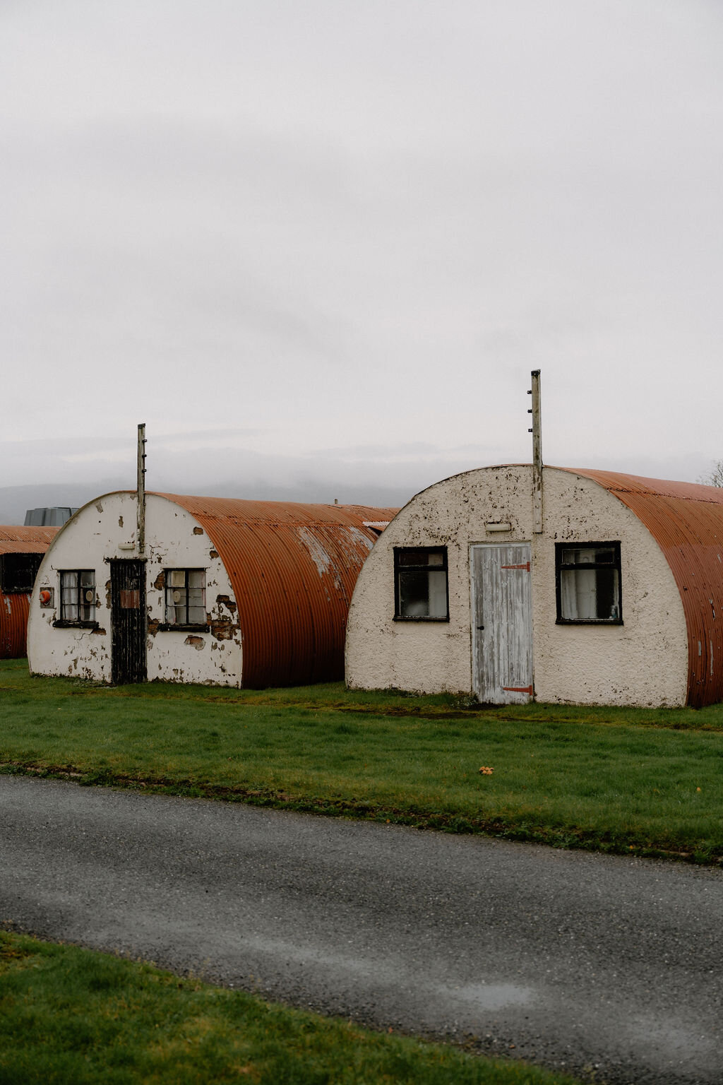 A rusted and a whitewashed Nissen hut sit side by side on the grass beside a road.
