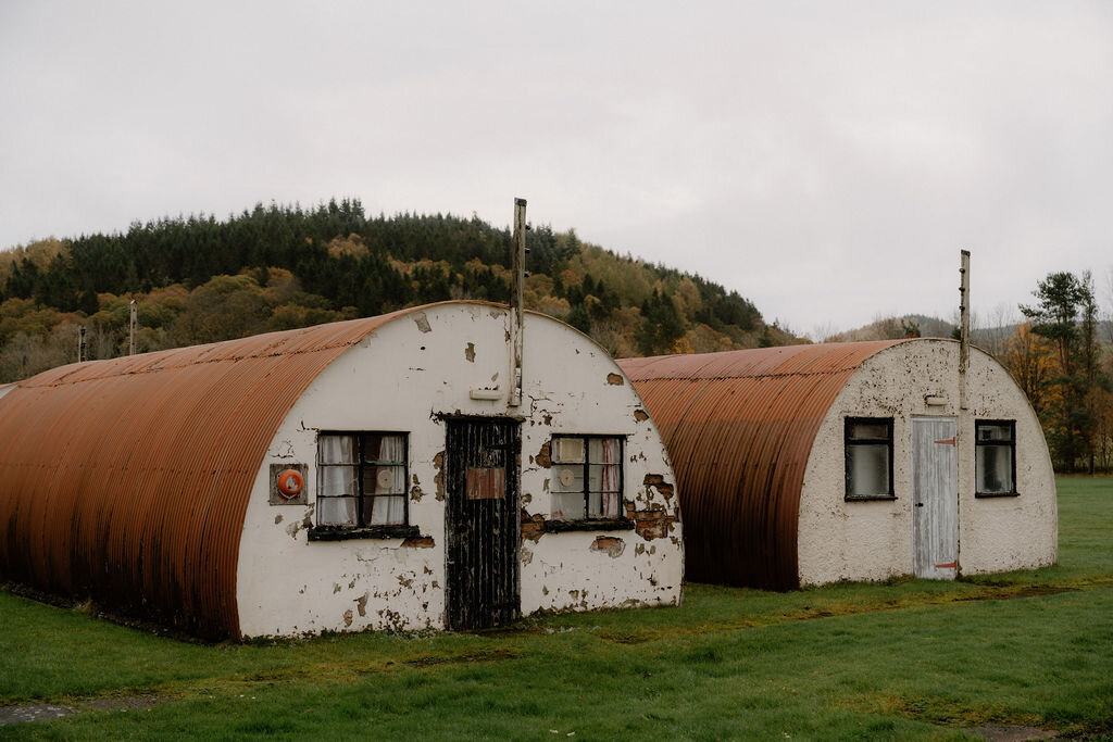 A rusted and a whitewashed Nissen hut stand beside each other before autumn-coloured hills.
