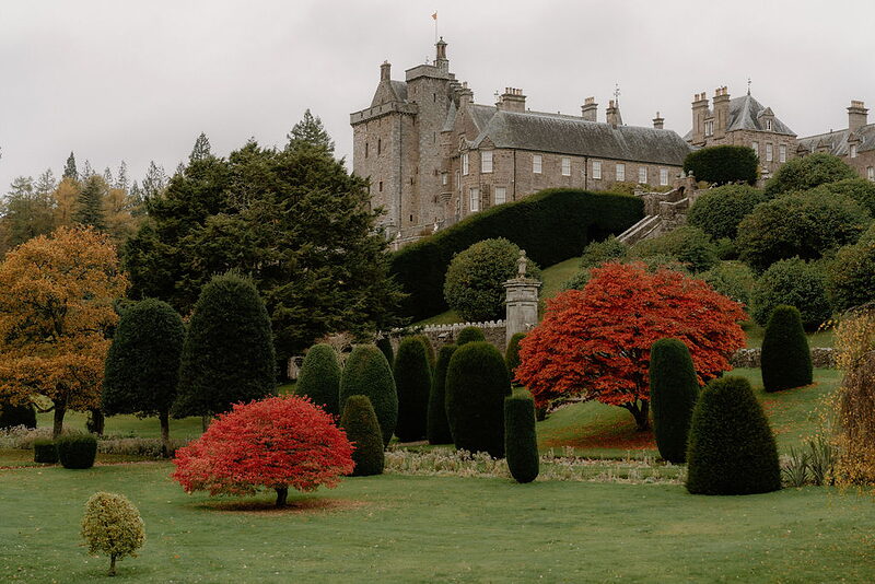 Red Japanese maples punctuate a clipped topiary garden before a turreted mansion.