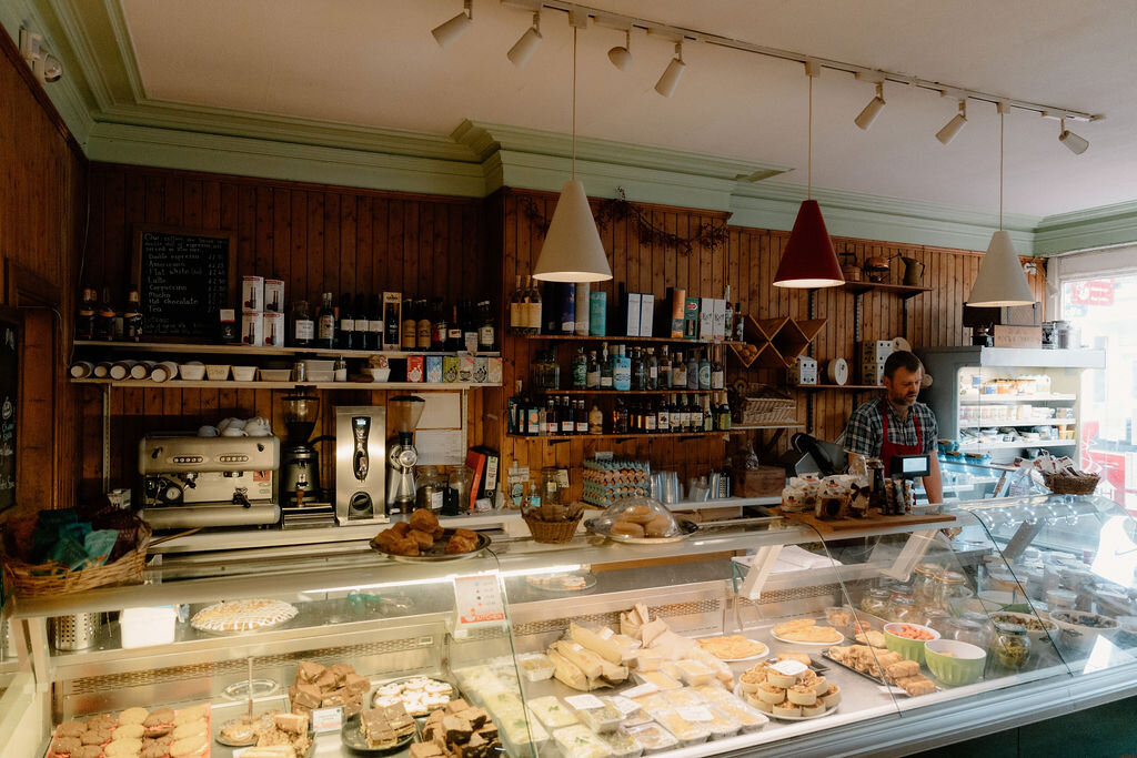A shopkeeper works behind a deli counter stocked with cheeses and prepared foods beneath pendant lights.
