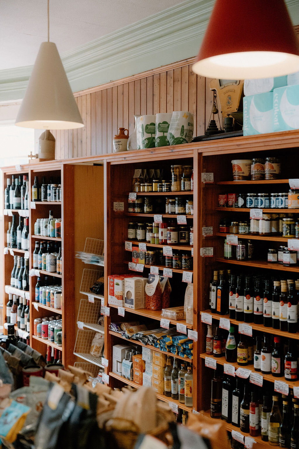 Wine bottles, jars and jams fill a shop's wooden shelving beneath pendant lights.