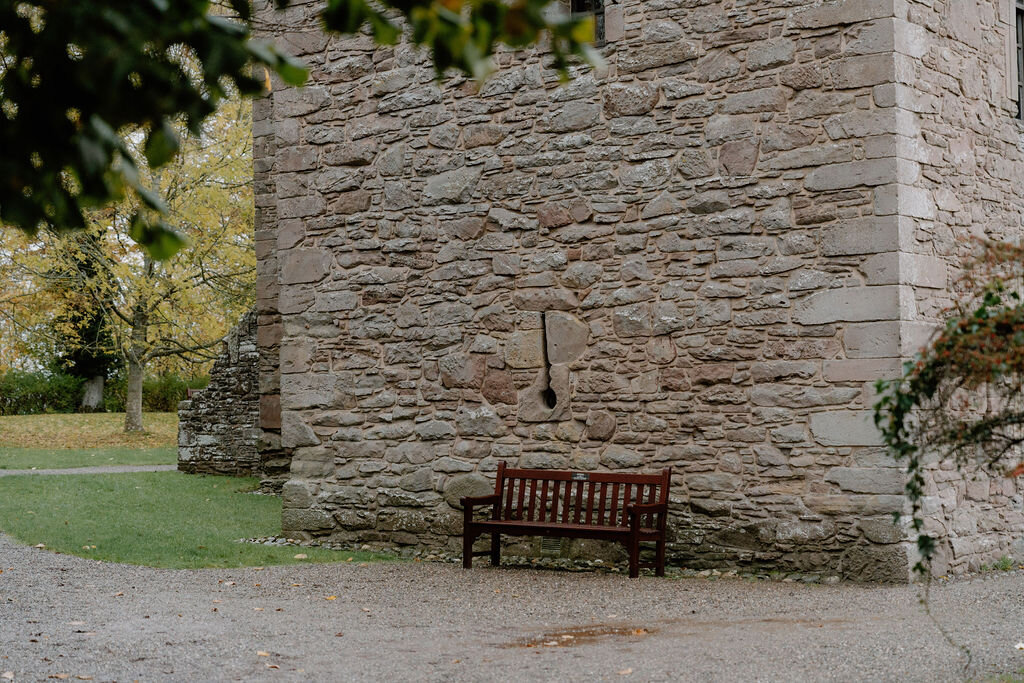 A wooden bench sits against a castle's stone wall beneath autumn trees.