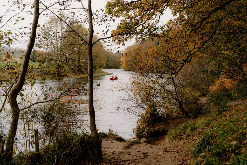 Two people paddling a red canoe along the river at Wildwoodz, autumn trees reflected in the water.