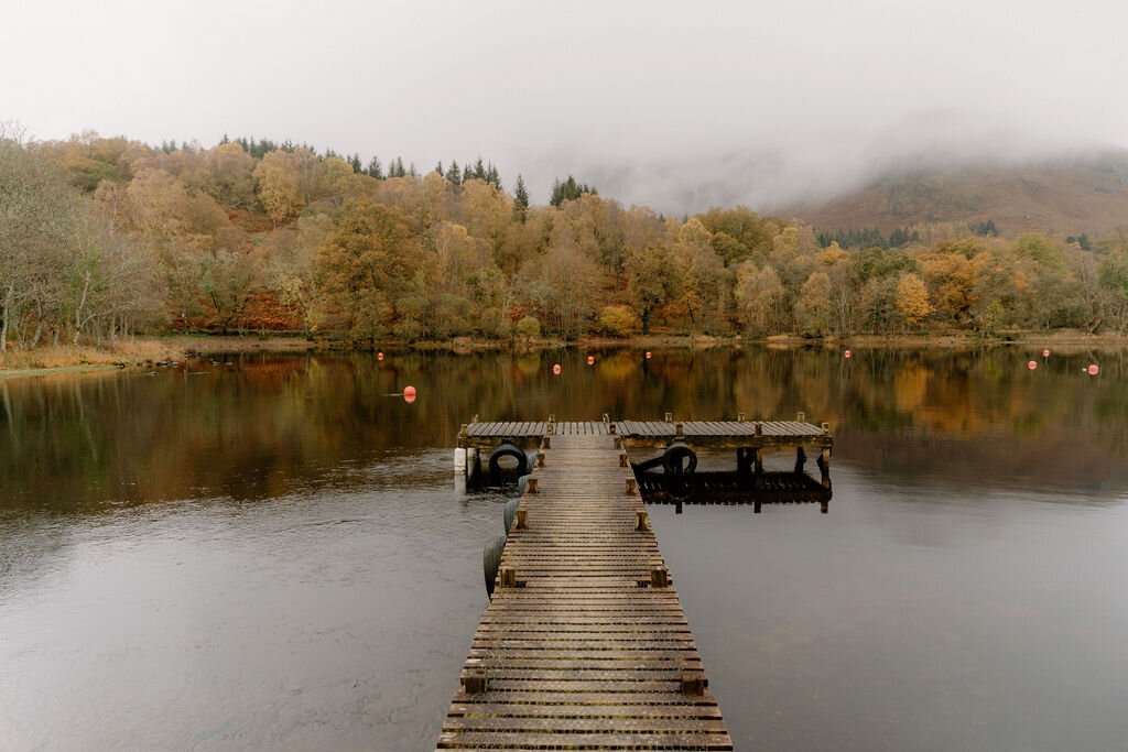 A wooden jetty with mooring pontoons extends into a still loch near Wildwoodz, autumn-coloured woodland reflected in the water.