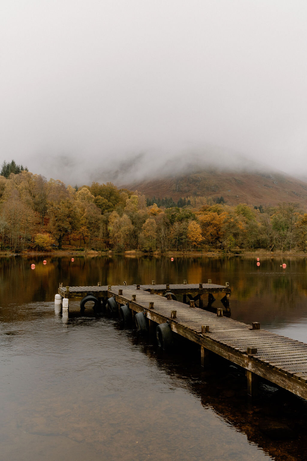 A wooden jetty stretches into a still loch near Wildwoodz, autumn trees fading into mist beneath a cloud-covered hillside.