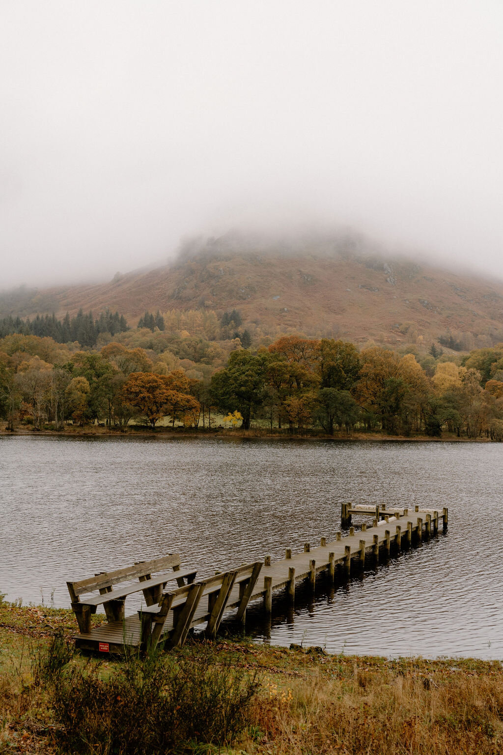 A wooden jetty with benches extends into a calm loch near Wildwoodz, a mist-covered hill rising above autumn trees on the far shore.