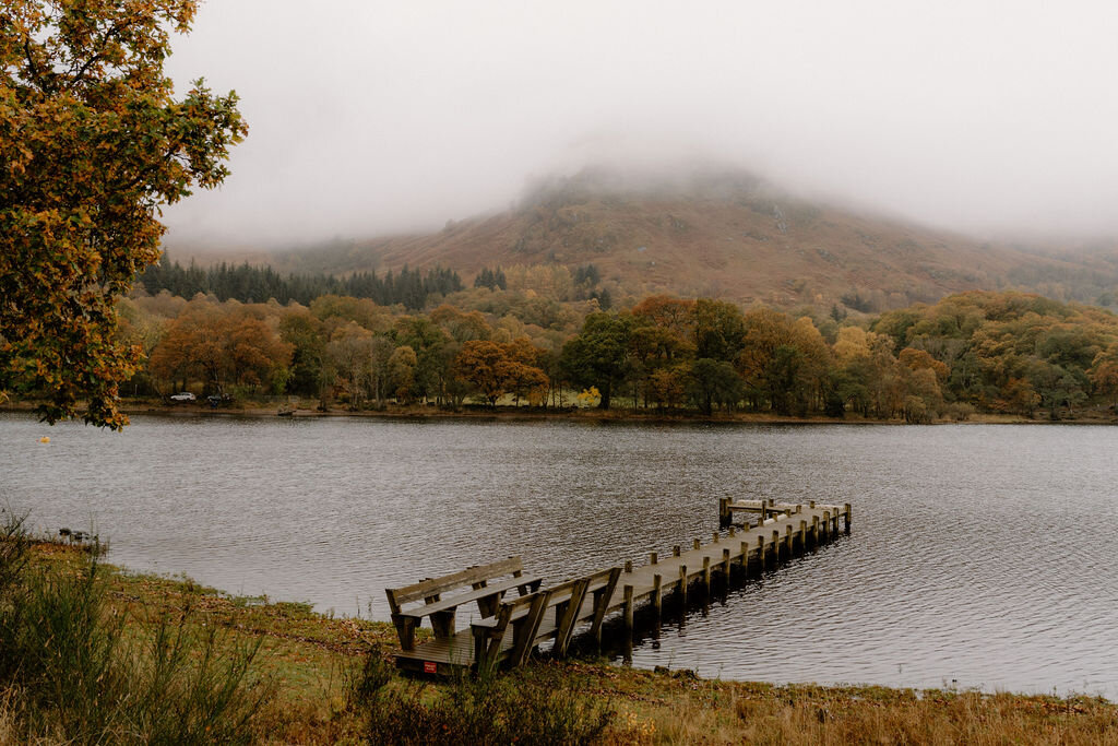 A wooden jetty extends into a calm loch near Wildwoodz, a mist-covered hill rising above autumn-coloured trees on the far shore.
