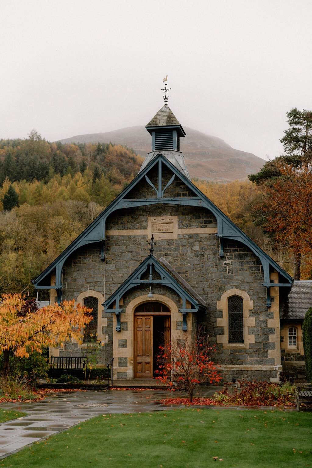 A small stone church with blue-painted eaves and a bell turret stands among autumn trees near Wildwoodz, a weathervane topping its spire.