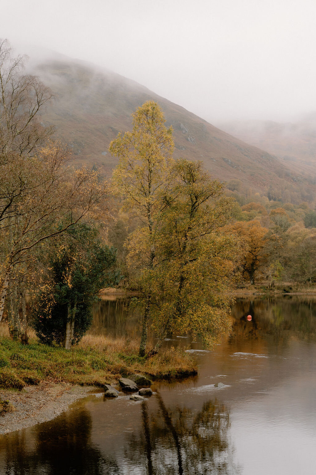 Autumn-coloured trees line the shore of a still loch near Wildwoodz, a red buoy floating near rocks at the water's edge.