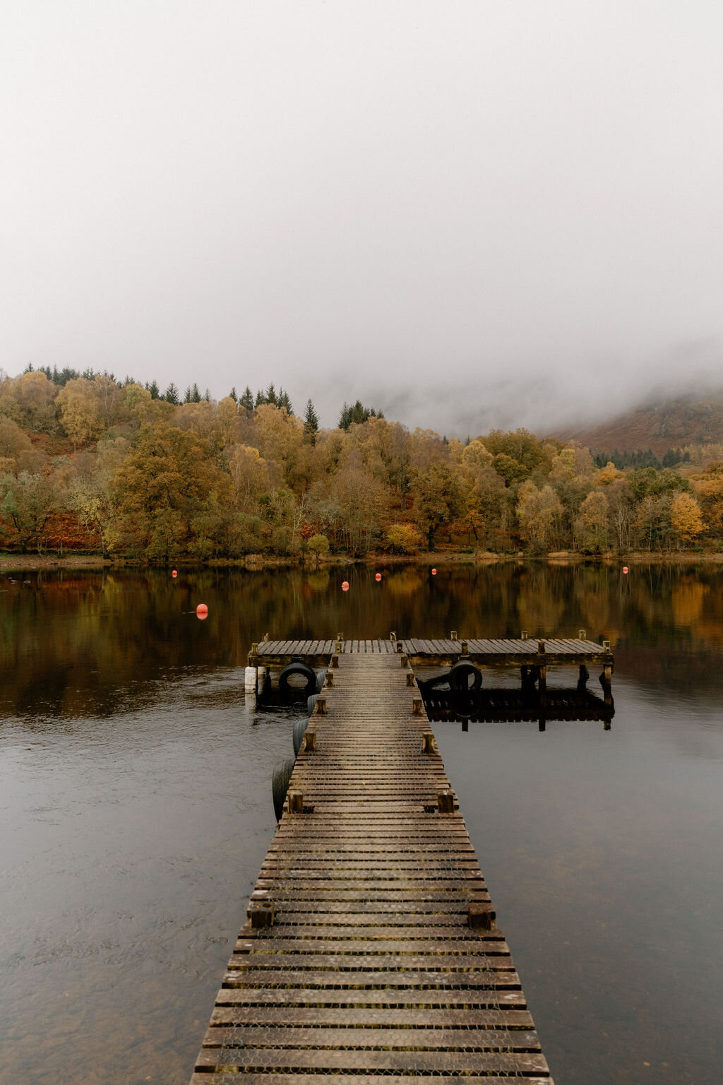 A wooden jetty with mooring tyres extends into a misty loch near Wildwoodz, autumn-coloured trees reflected in the still water.